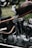 A close-up view of a custom motorcycle, showcasing intricate detailing on the gas tank and a unique, curved metal kickstand. The engine components are prominently displayed, highlighting the craftsmanship and vintage style.