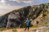 A person wearing outdoor gear stands on a grassy ledge, overlooking a rugged mountain landscape. The mountains are covered with dense green pine trees and rocky cliffs illuminated by sunlight. Clouds drift across the sky, adding a serene backdrop to the scene.