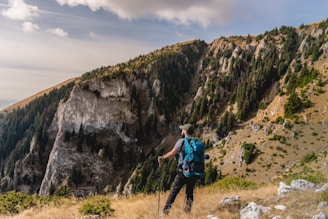 A person wearing outdoor gear stands on a grassy ledge, overlooking a rugged mountain landscape. The mountains are covered with dense green pine trees and rocky cliffs illuminated by sunlight. Clouds drift across the sky, adding a serene backdrop to the scene.