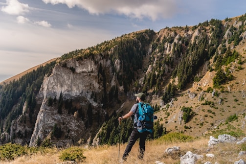 A person wearing outdoor gear stands on a grassy ledge, overlooking a rugged mountain landscape. The mountains are covered with dense green pine trees and rocky cliffs illuminated by sunlight. Clouds drift across the sky, adding a serene backdrop to the scene.