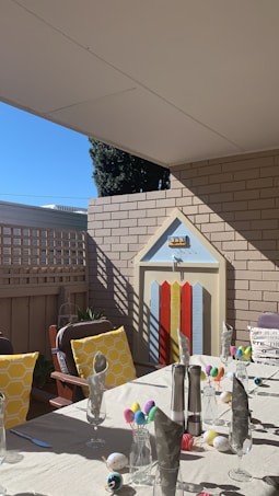A patio area set up for a festive gathering, featuring a table adorned with colorful decorations, including vibrant eggs in glasses. The table is covered with a beige cloth, and there are napkins arranged pointedly. Bright yellow pillows with a honeycomb pattern are placed on wooden chairs. A playful door with a striped design is set into a brick wall at the back, adding a whimsical touch. The clear blue sky and sunlight enhance the cheerful atmosphere.