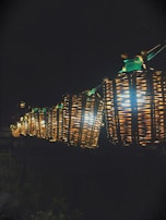 A row of glowing wicker lanterns suspended in the air, illuminating the surrounding darkness with a warm, inviting light. The lanterns are intricately woven, displaying a rustic and handcrafted aesthetic. The nighttime setting enhances their brightness and creates a cozy ambiance.