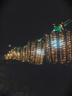 A row of glowing wicker lanterns suspended in the air, illuminating the surrounding darkness with a warm, inviting light. The lanterns are intricately woven, displaying a rustic and handcrafted aesthetic. The nighttime setting enhances their brightness and creates a cozy ambiance.