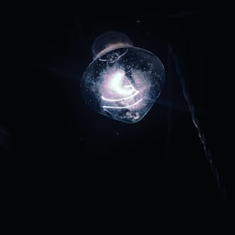 An illuminated light bulb glowing in the dark. The bulb appears slightly dusty or dirty, emitting a soft, bluish-white light. The surrounding area is dark, with a faint outline of a coiled cord or wire visible on the right.