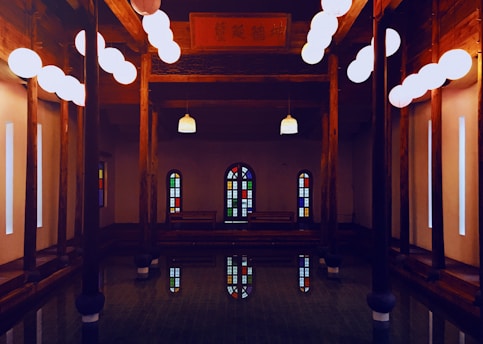 A serene indoor environment features wooden columns and warm lighting from spherical hanging lamps. Stained glass windows add colorful reflections to the tranquil water surface below, creating a peaceful and contemplative atmosphere.