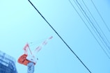 A construction crane stands prominently against a clear blue sky, with power lines running diagonally across the image. The crane's tower and boom are painted in bright red and white colors. Part of a scaffolding-covered building is visible on the left side of the frame.