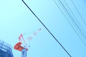 A construction crane stands prominently against a clear blue sky, with power lines running diagonally across the image. The crane's tower and boom are painted in bright red and white colors. Part of a scaffolding-covered building is visible on the left side of the frame.