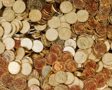 A large pile of mixed coins, predominantly of Australian origin, with varying shades of gold and copper. The coins are densely packed and form a textured, reflective surface.