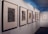 A neatly arranged display of framed artworks is mounted on a white wall within what appears to be an art gallery. The frames are predominantly wooden, and the artworks within range from abstract to detailed black and white sketches or prints. The gallery has soft lighting that highlights the artwork and a reflective ceiling, adding a modern touch to the space.