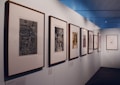 A neatly arranged display of framed artworks is mounted on a white wall within what appears to be an art gallery. The frames are predominantly wooden, and the artworks within range from abstract to detailed black and white sketches or prints. The gallery has soft lighting that highlights the artwork and a reflective ceiling, adding a modern touch to the space.