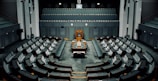 A large, empty parliamentary chamber with tiered seating arranged in a semicircular formation around a central podium. The room features muted colors, primarily gray and dark teal, with a classic design incorporating wooden elements. There are multiple levels of seating which face the main desk positioned under a large clock.