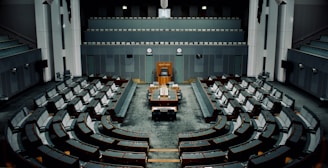 A large, empty parliamentary chamber with tiered seating arranged in a semicircular formation around a central podium. The room features muted colors, primarily gray and dark teal, with a classic design incorporating wooden elements. There are multiple levels of seating which face the main desk positioned under a large clock.