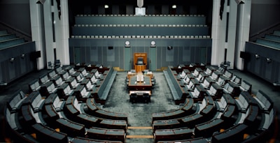 A large, empty parliamentary chamber with tiered seating arranged in a semicircular formation around a central podium. The room features muted colors, primarily gray and dark teal, with a classic design incorporating wooden elements. There are multiple levels of seating which face the main desk positioned under a large clock.