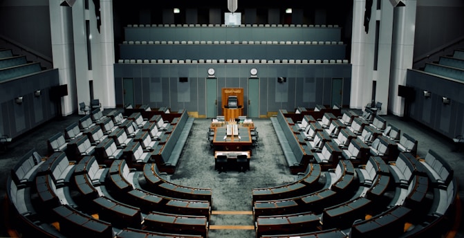 A large, empty parliamentary chamber with tiered seating arranged in a semicircular formation around a central podium. The room features muted colors, primarily gray and dark teal, with a classic design incorporating wooden elements. There are multiple levels of seating which face the main desk positioned under a large clock.