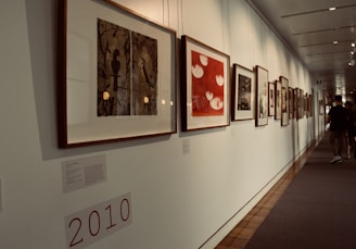 A well-lit art gallery hallway featuring a series of framed artworks hung on a white wall. The artworks vary in style and color, with some pieces displaying abstract patterns and others showcasing more figurative elements. The floor is a blend of carpet and wood, and a sign with '2010' is visible. People are casually walking, indicating movement and interaction within the space.