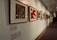 A well-lit art gallery hallway featuring a series of framed artworks hung on a white wall. The artworks vary in style and color, with some pieces displaying abstract patterns and others showcasing more figurative elements. The floor is a blend of carpet and wood, and a sign with '2010' is visible. People are casually walking, indicating movement and interaction within the space.