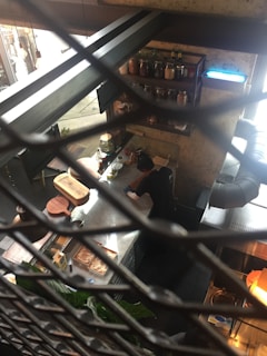 A kitchen environment is viewed from above, enclosed with a metal mesh. A person wearing dark clothing is seen working at a stainless steel countertop, surrounded by various kitchen tools and ingredients. Shelves are lined with glass jars and bottles, and there is a hint of natural light coming in from the window.