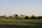 A rural landscape featuring a series of farm buildings, including a barn and several other agricultural structures, surrounded by lush green fields. The horizon shows clear skies and a few trees scattered around the premises.