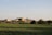 A rural landscape featuring a series of farm buildings, including a barn and several other agricultural structures, surrounded by lush green fields. The horizon shows clear skies and a few trees scattered around the premises.