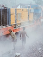 Two individuals stand next to a large industrial machine, surrounded by a thick haze of dust. The machine is yellow with various vents and labeled 'XRV 1200'. The ground is rough and covered in debris, suggesting a construction or work site. Both individuals are dressed in workwear, and visibility is low due to the dust in the air.