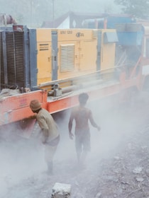 Two individuals stand next to a large industrial machine, surrounded by a thick haze of dust. The machine is yellow with various vents and labeled 'XRV 1200'. The ground is rough and covered in debris, suggesting a construction or work site. Both individuals are dressed in workwear, and visibility is low due to the dust in the air.