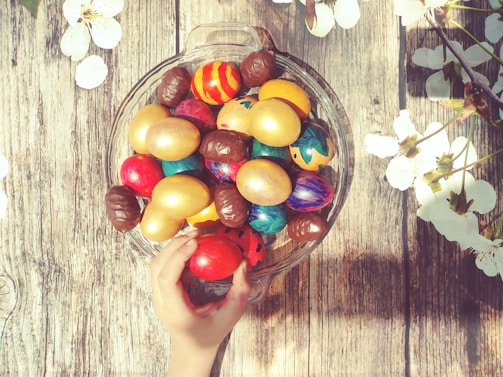 A collection of brightly colored, decorated eggs and golden eggs inside a glass bowl placed on a rustic wooden surface. A hand is reaching for an egg, surrounded by delicate white flowers that are scattered upon the wooden background, adding a touch of nature.