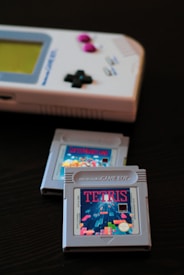 A classic Nintendo Game Boy with its distinctive grey color and control buttons next to two Game Boy cartridges. One cartridge is prominently displaying the Tetris label, while the background cartridge is barely visible.