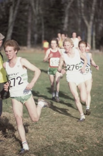 A group of male runners are participating in an outdoor race, each wearing numbered bibs, shorts, and running shoes. They are running on a grassy field with trees in the background on a clear day.