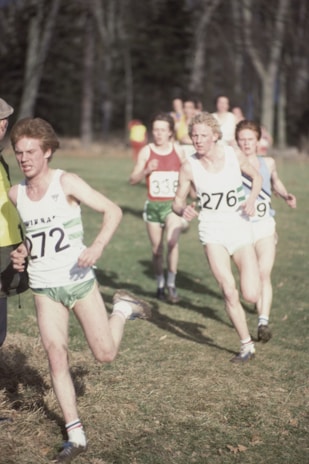 A group of male runners are participating in an outdoor race, each wearing numbered bibs, shorts, and running shoes. They are running on a grassy field with trees in the background on a clear day.