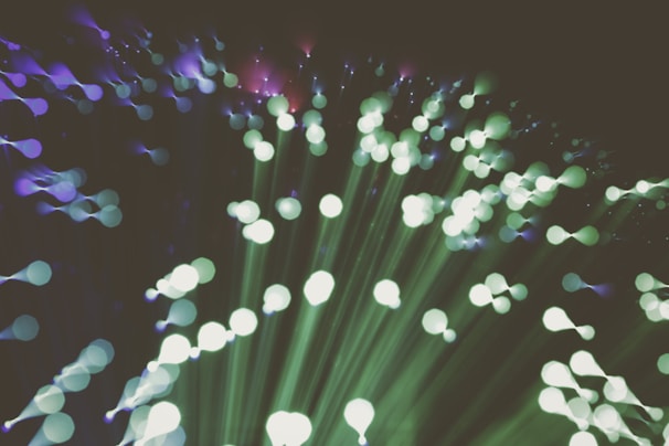 A captivating abstract representation of fiber optic lights, featuring an array of radiant, out-of-focus circles in green and purple hues that create an ethereal, flowing pattern. The lights appear to burst outward, creating a sense of movement and expansion.