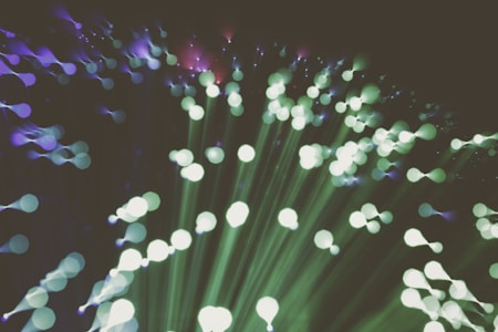 A captivating abstract representation of fiber optic lights, featuring an array of radiant, out-of-focus circles in green and purple hues that create an ethereal, flowing pattern. The lights appear to burst outward, creating a sense of movement and expansion.