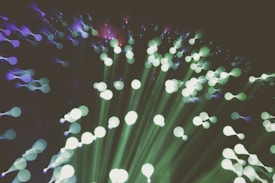 A captivating abstract representation of fiber optic lights, featuring an array of radiant, out-of-focus circles in green and purple hues that create an ethereal, flowing pattern. The lights appear to burst outward, creating a sense of movement and expansion.