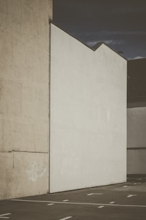 A minimalistic urban scene featuring a large blank white wall and a adjacent beige wall with some faint graffiti near the bottom. The concrete ground in the foreground has white painted lines, indicating a parking area. The sky above appears to be overcast or darkening, adding a somber tone to the image.
