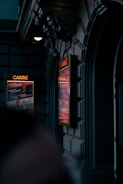 A dimly lit exterior of a theater entrance with ornate architectural details and two illuminated posters displaying events or performances. The dark stone facade and decorative sculptures add an elegant touch to the setting.