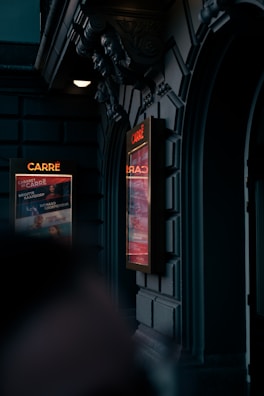 A dimly lit exterior of a theater entrance with ornate architectural details and two illuminated posters displaying events or performances. The dark stone facade and decorative sculptures add an elegant touch to the setting.