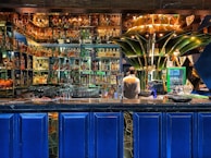 A vibrant bar setting filled with an extensive collection of liquor bottles arranged on illuminated shelves. A bartender is standing behind the counter, seemingly preparing drinks. The decor includes modern, artistic elements, with a prominent green structure at the center and deep blue panels below the counter.