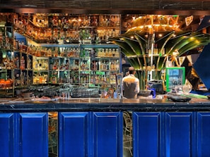 A vibrant bar setting filled with an extensive collection of liquor bottles arranged on illuminated shelves. A bartender is standing behind the counter, seemingly preparing drinks. The decor includes modern, artistic elements, with a prominent green structure at the center and deep blue panels below the counter.
