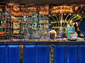 A vibrant bar setting filled with an extensive collection of liquor bottles arranged on illuminated shelves. A bartender is standing behind the counter, seemingly preparing drinks. The decor includes modern, artistic elements, with a prominent green structure at the center and deep blue panels below the counter.