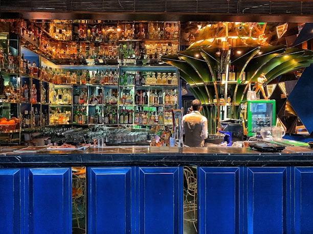 A vibrant bar setting filled with an extensive collection of liquor bottles arranged on illuminated shelves. A bartender is standing behind the counter, seemingly preparing drinks. The decor includes modern, artistic elements, with a prominent green structure at the center and deep blue panels below the counter.