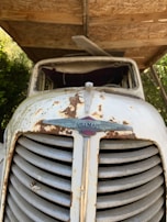 A vintage Hillman vehicle front view displays significant rust and wear on its grill and body. The emblem and brand name are visible amidst the corroded and discolored metal surfaces. The vehicle is situated under a wooden shelter with greenery visible in the background.