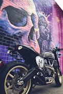 A black motorcycle with gold rims is parked in front of a colorful brick wall mural. The mural features a large and abstract depiction of a face or skull in vibrant purples, blues, and pinks. The motorcycle has a number 54 painted on its side.
