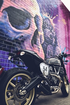 A black motorcycle with gold rims is parked in front of a colorful brick wall mural. The mural features a large and abstract depiction of a face or skull in vibrant purples, blues, and pinks. The motorcycle has a number 54 painted on its side.