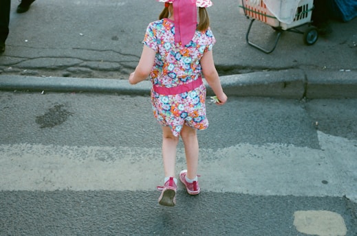 A young child in a colorful floral outfit and pink hat walks across a street. The child is wearing pink shoes and is seen from the back. There is a cart with wheels in the background and part of another person's leg is visible on the left.