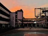 An outdoor basketball court is surrounded by tall buildings with a vibrant orange sky at dusk. A basketball hoop is prominently visible on the right, while several people and a dog are seen walking on the court.