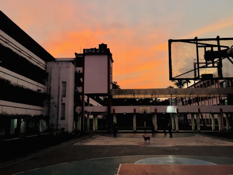 An outdoor basketball court is surrounded by tall buildings with a vibrant orange sky at dusk. A basketball hoop is prominently visible on the right, while several people and a dog are seen walking on the court.