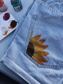 A pair of light blue denim jeans with a hand-painted sunflower design on one of the pockets. Next to the jeans are several small bottles of paint, including orange, green, and purple. A small palette with paint smudges is also visible.