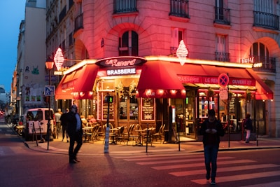 A street corner at dusk with a warmly lit brasserie featuring red awnings and neon signs. People are seated at outdoor tables, and pedestrians are walking by, creating a lively and inviting atmosphere. The brasserie has a traditional look with decorative lighting enhancing the welcoming vibe.