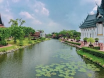 A serene landscape featuring a tranquil river flanked by lush greenery and traditional architectural structures. The river is adorned with lily pads, and the banks are lined with neatly trimmed bushes and ornate benches, showcasing a blend of nature and elegant design.