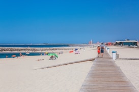 A sandy beach with several umbrellas and people relaxing near the water. A wooden boardwalk stretches across the sand, leading towards a lighthouse and buildings in the distance. A couple walks along the boardwalk, with a vibrant blue sky overhead.