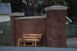 A wooden bench with the words 'WATCH LISTEN REFLECT' painted on it is placed against a tall red brick wall between two pillars. The setting includes a chain link fence with some foliage in the background, and a house partially visible in the distance.